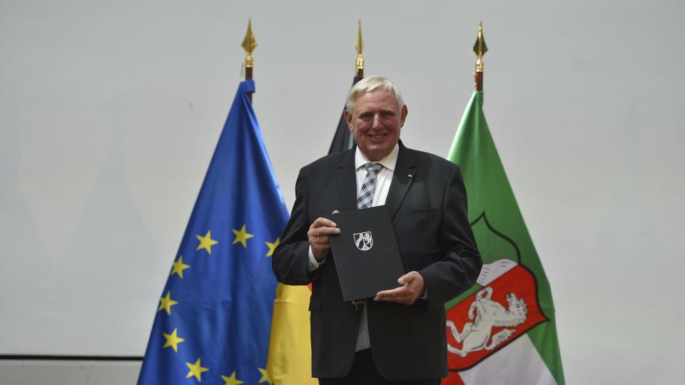 Minister Laumann steht mit seiner Urkaunde zur Ernennung als Minister vor drei Fahnen