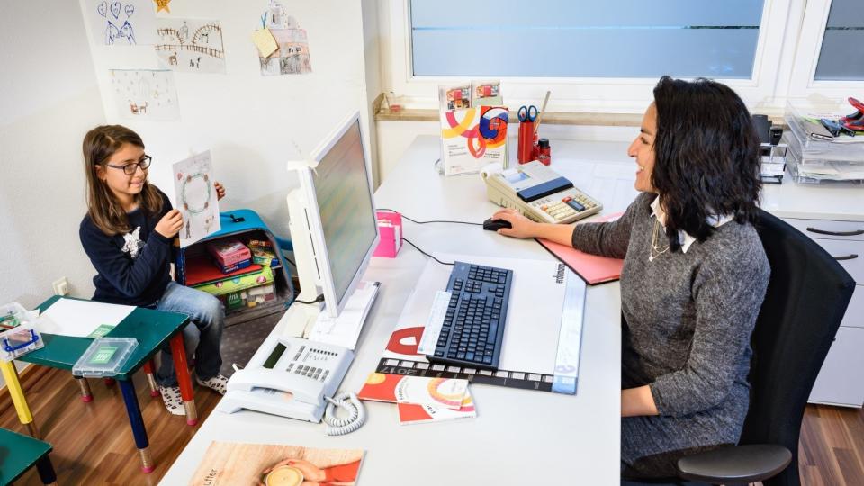 Foto zeigt Auszubildende an ihrem Computerarbeitsplatz und ihre Tochter in einer Spielecke