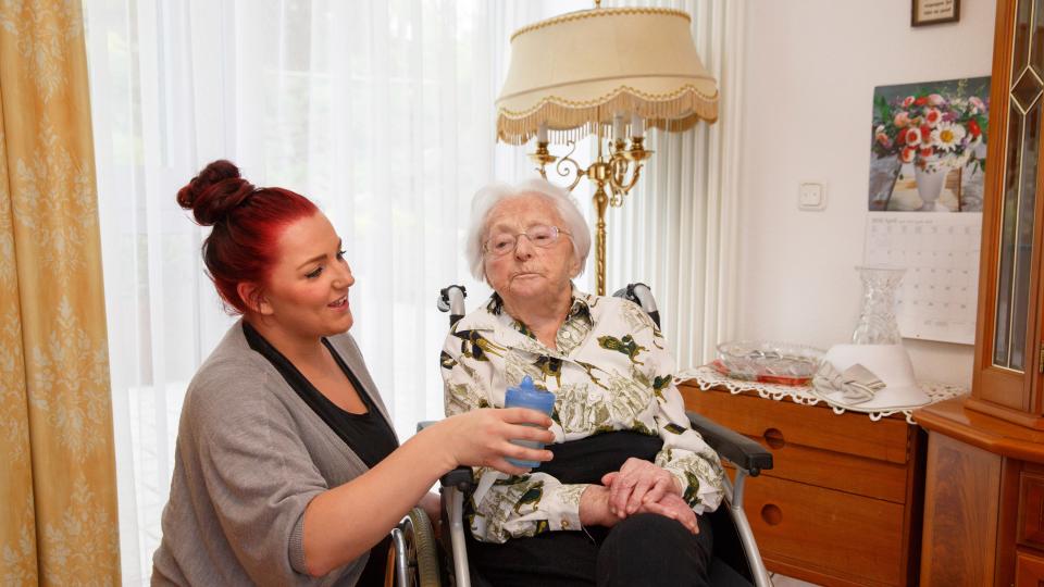 Foto: Junge Frau bietet einer älteren Damen, die im Rollstuhl sitzt, einen Becher zum Trinken