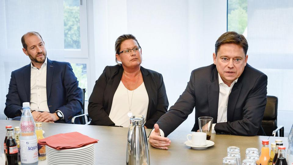 Zwei Männer und eine Frau sitzen am Tisch, der Mann rechts hält das Wort