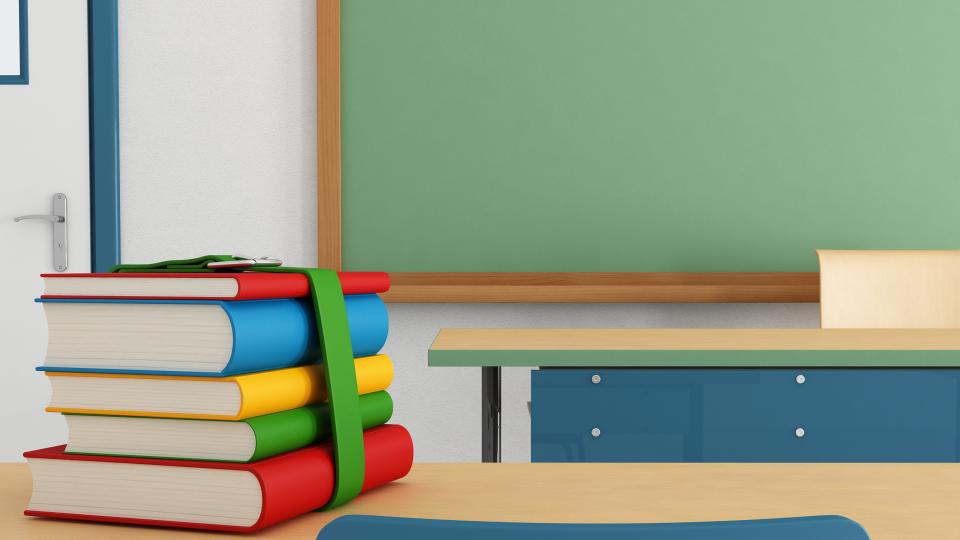 Foto zeigt Schulbücher auf einem Tisch vor einer Tafel im Klassenzimmer