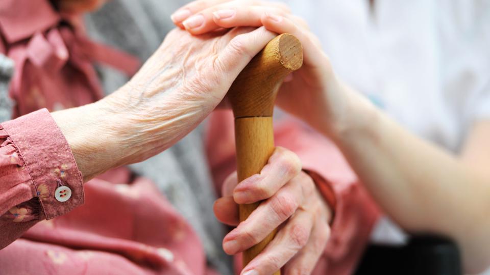 Foto zeigt pfegebedürftige Frau mit Hand an einem Gehstock, auf der die Hand einer Helferin liegt