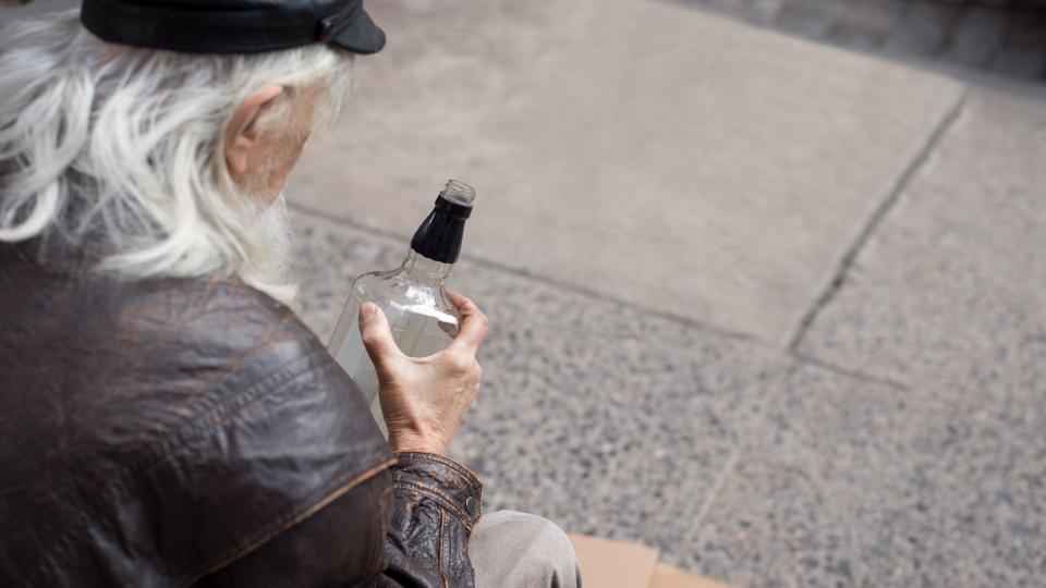 Alter Mann sitzt auf der Straße und blickt auf eine Flasche in seiner Hand
