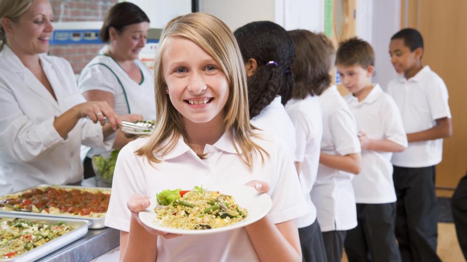 Lächendes Mädchen hält ihr Mittagessenteller in die Kamera. Im Hintergrund sind Kinder in der Schlange zur Essensausgabe.