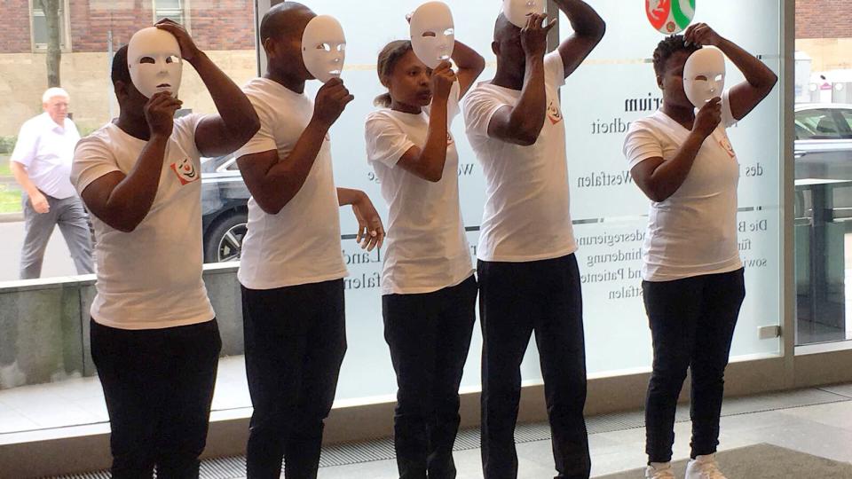 Fünf Mitglieder der Pantomime-Gruppe „Mime Art for Life“ stehen nebeneinander und setzen weiße Masken auf.